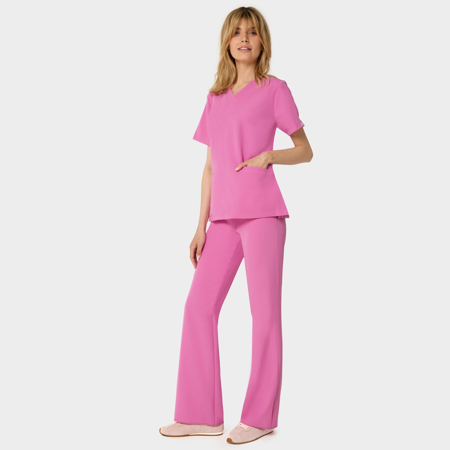 Bluza medyczna Emily w kolorze Disco Pink od Vena Uniformy.
Modelka stoi na jasnym tle w komplecie medycznym: bluza z krótkim rękawem oraz spodnie flare, zestawione z beżowymi sneakersami.