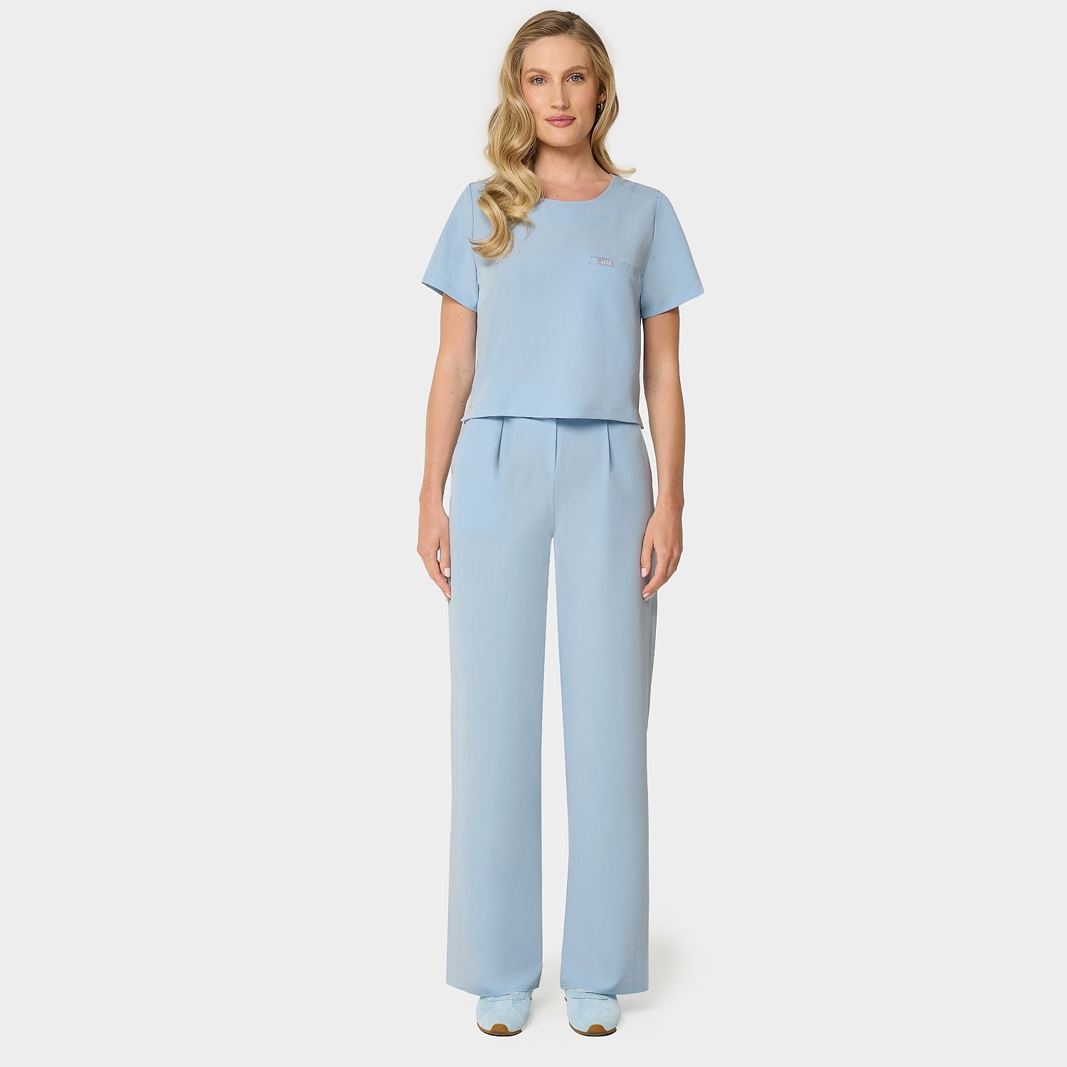 Bluza medyczna Gaja crop w kolorze BABY BLUE od Vena Uniformy.
Modelka stoi na jasnym tle w pastelowym komplecie medycznym: bluza z krótkim rękawem oraz szerokie spodnie, zestawione z niebieskimi sneakersami.