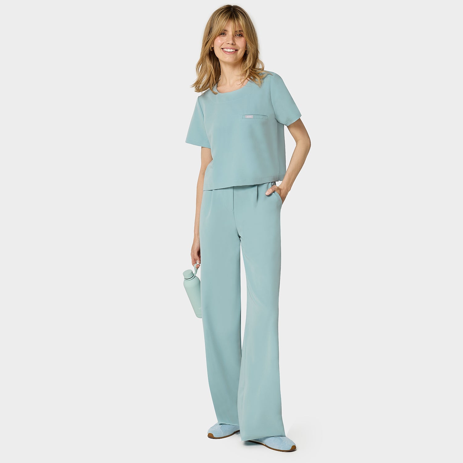 Bluza medyczna Gaja crop w kolorze CLOUDY BLUE od Vena Uniformy.
Modelka stoi na jasnym tle w pastelowym komplecie medycznym: bluza z krótkim rękawem oraz szerokie spodnie, zestawione z niebieskimi sneakersami.