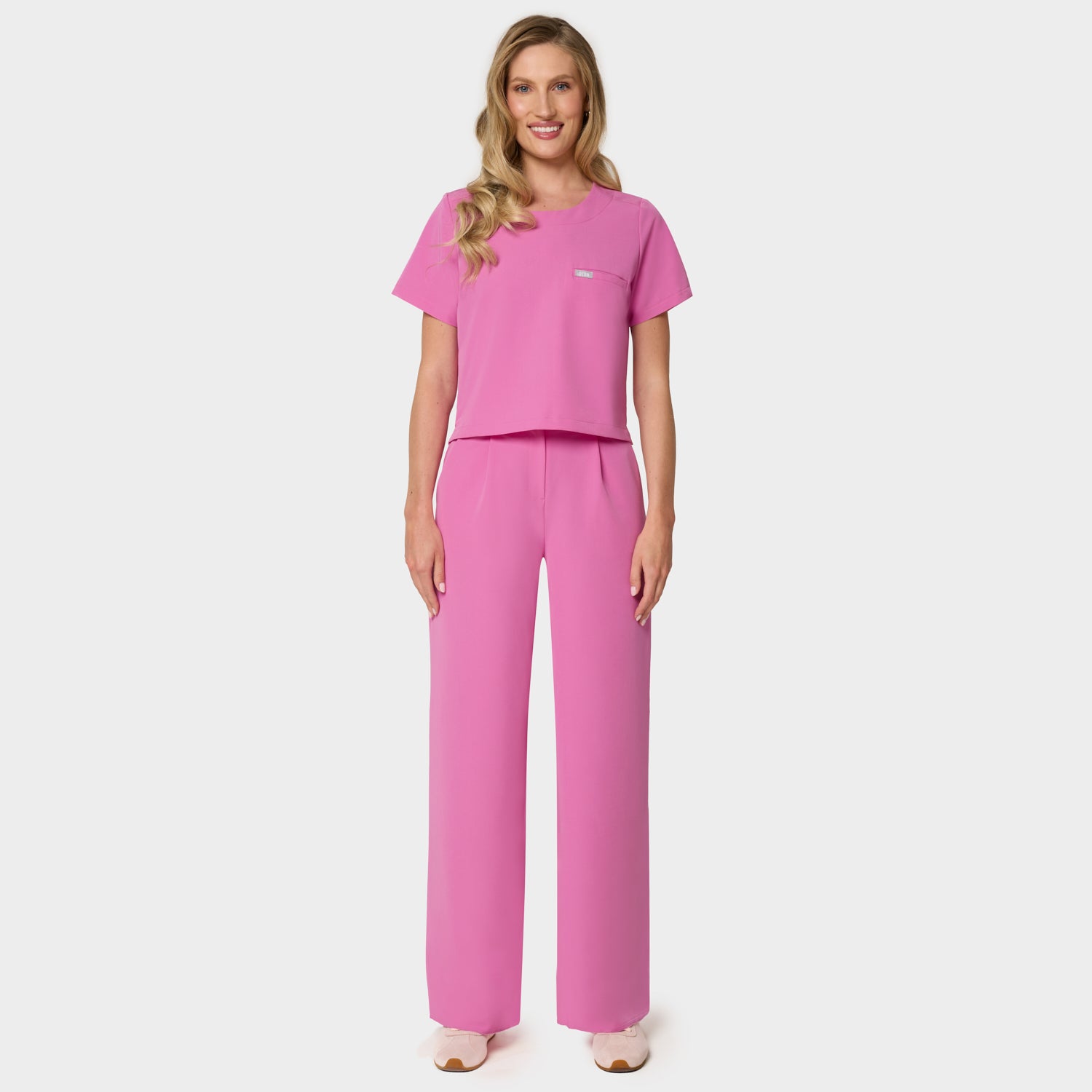 Bluza medyczna Gaja crop w kolorze Disco Pink od Vena Uniformy.
Modelka stoi na jasnym tle w komplecie medycznym: bluza z krótkim rękawem oraz szerokie spodnie, zestawione z beżowymi sneakersami.