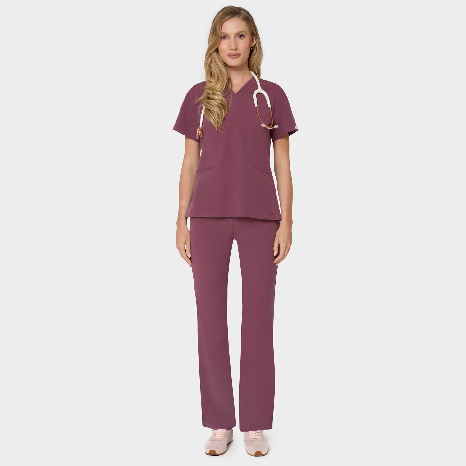 Bluza medyczna Emily w kolorze Dusty Plum od Vena Uniformy.
Modelka stoi na jasnym tle w komplecie medycznym: bluza z krótkim rękawem oraz spodnie flare, zestawione z beżowymi sneakersami.