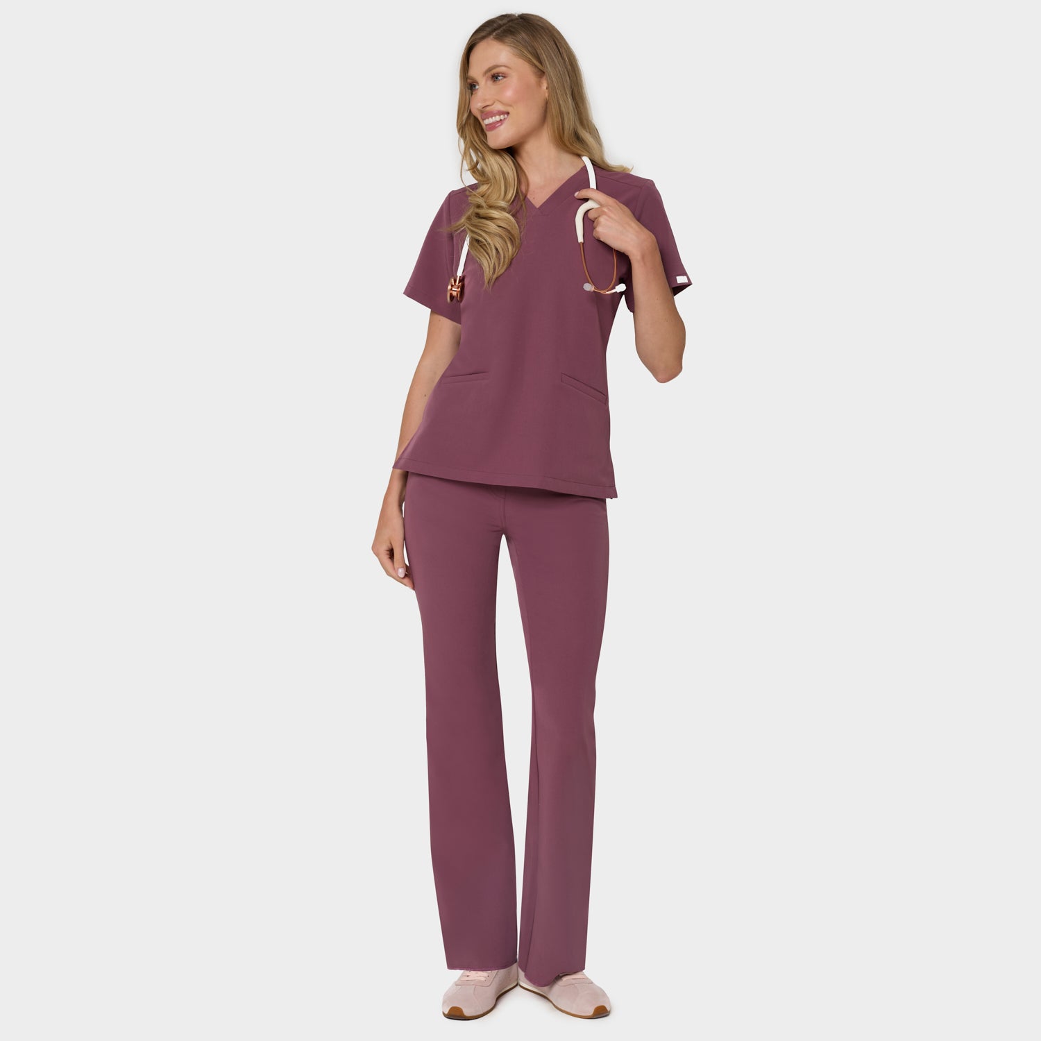 Bluza medyczna Emily w kolorze Dusty Plum od Vena Uniformy.
Modelka stoi na jasnym tle w komplecie medycznym: bluza z krótkim rękawem oraz spodnie flare, zestawione z beżowymi sneakersami.
