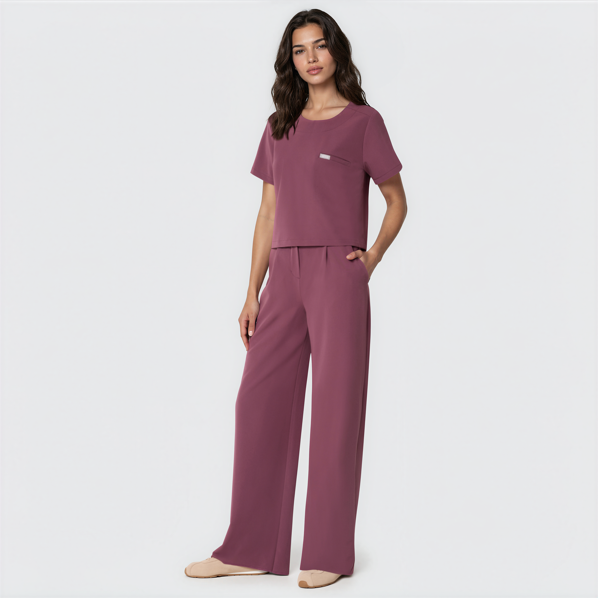 Bluza medyczna Gaja crop w kolorze Dusty Plum od Vena Uniformy.
Modelka na jasnym tle w komplecie medycznym: bluza z krótkim rękawem oraz szerokie spodnie, zestawione z beżowymi sneakersami.