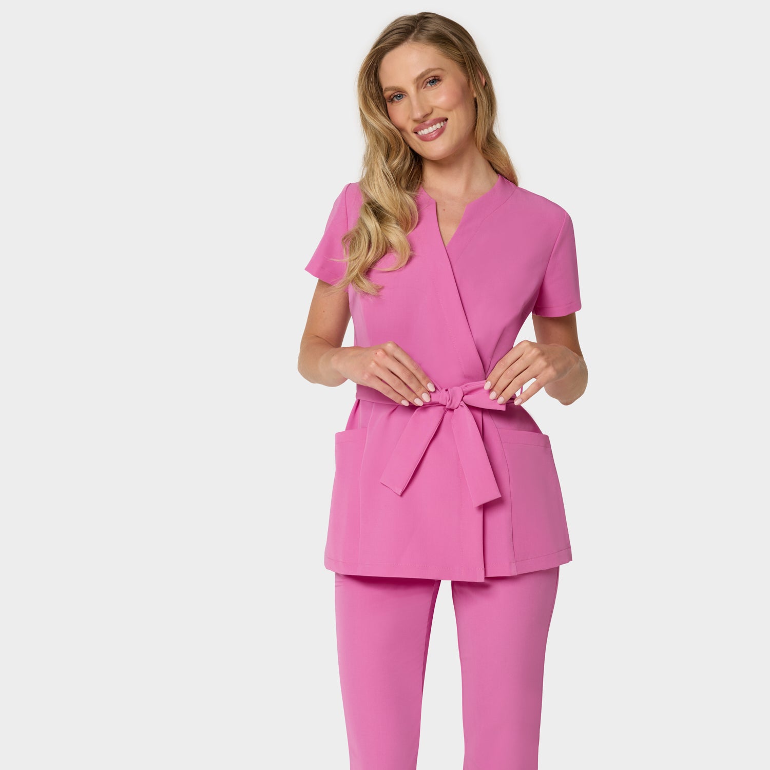 Kobieta w różowym fartuszku medycznym SOPHIA i dopasowanych spodniach flare — komplet w odcieniu Disco Pink, widok z przodu.